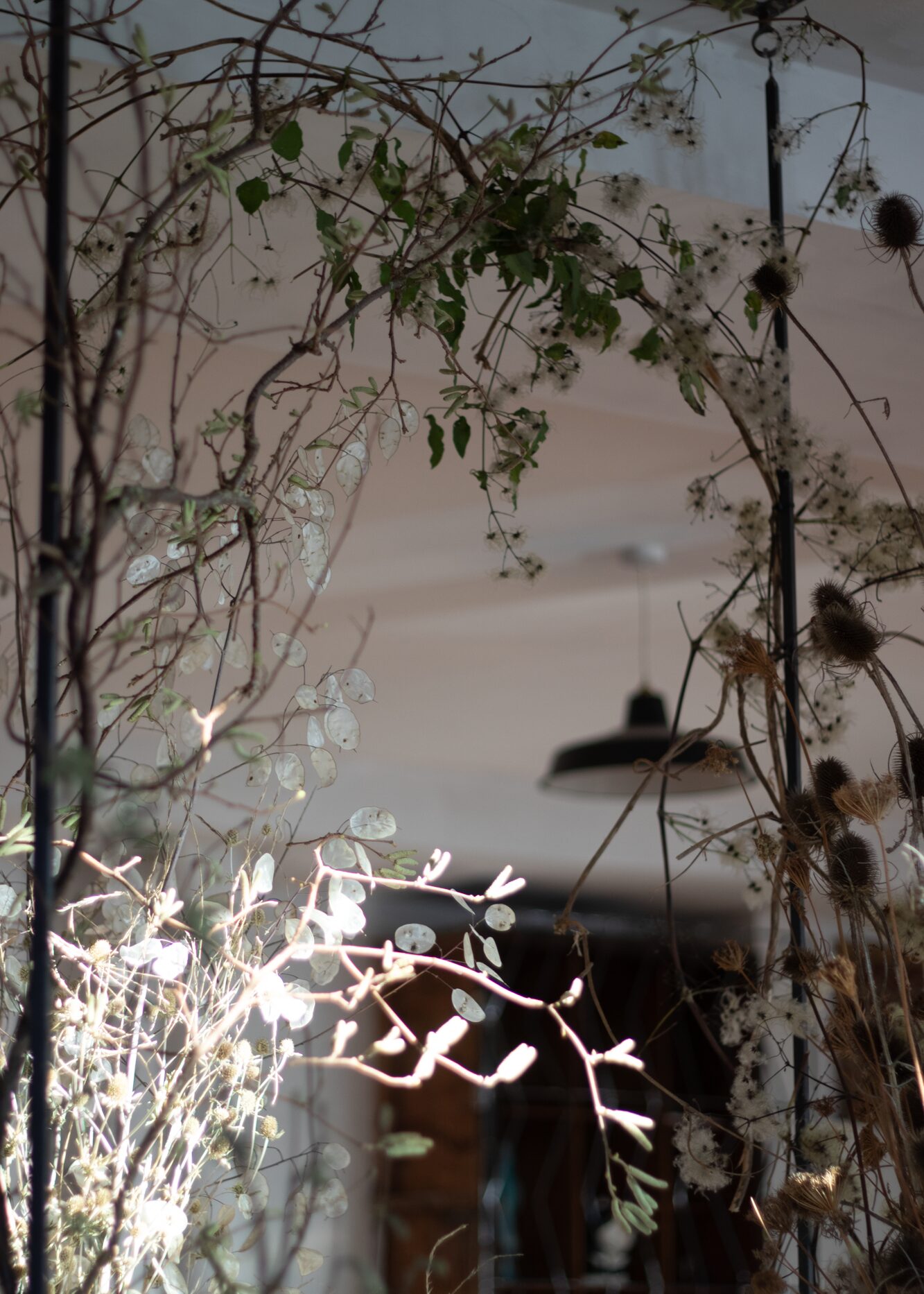 A sculptural winter scene. In the crisp light of the day, the layered display felt calm and airy. Once the sun had set, it would come to life. The tall teasels and skinny hazel branches would draw dramatic silhouettes and bold shadows, whilst the pearly lunaria discs and fluffy clematis seed heads would glow softly in the flickering candlelight. By Mora, a sustainable, season-led event florist based in Hackney, East London