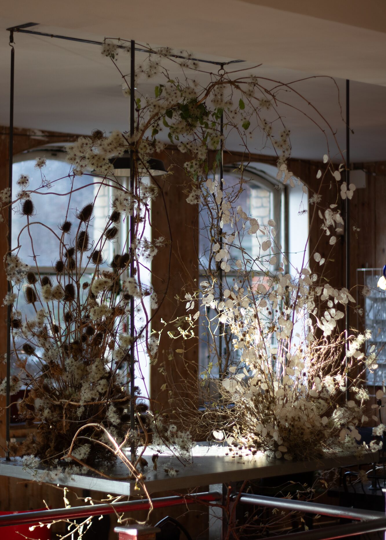 A sculptural winter scene. In the crisp light of the day, the layered display felt calm and airy. Once the sun had set, it would come to life. The tall teasels and skinny hazel branches would draw dramatic silhouettes and bold shadows, whilst the pearly lunaria discs and fluffy clematis seed heads would glow softly in the flickering candlelight. By Mora, a sustainable, season-led event florist based in Hackney, East London